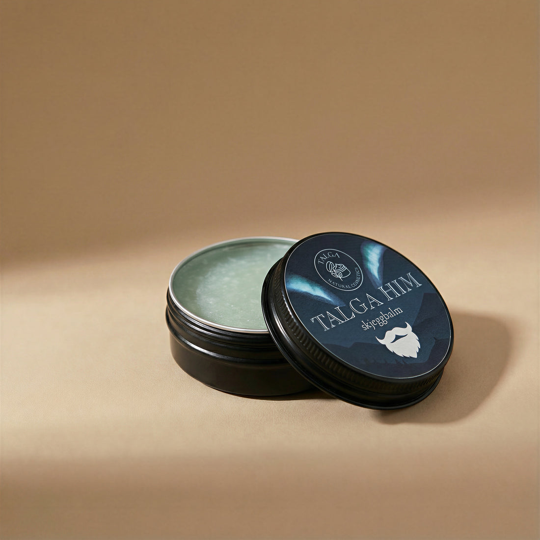 Open tin of Talga Him beard balm showing the balm surface, with the lid resting beside it on a beige background.
