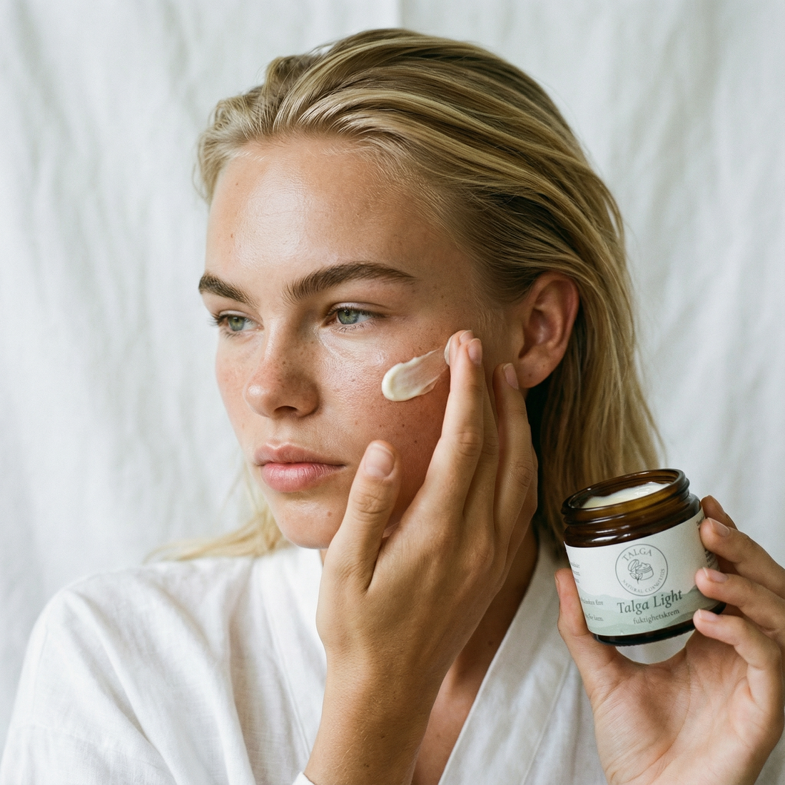 Woman applying Talga Light moisturizing cream to her cheek while holding the open jar.